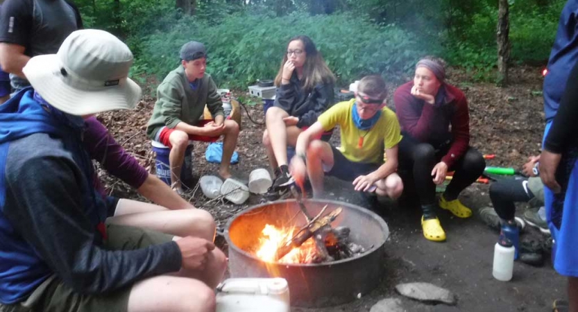 A group of people sit around a campfire in a wooded area. 